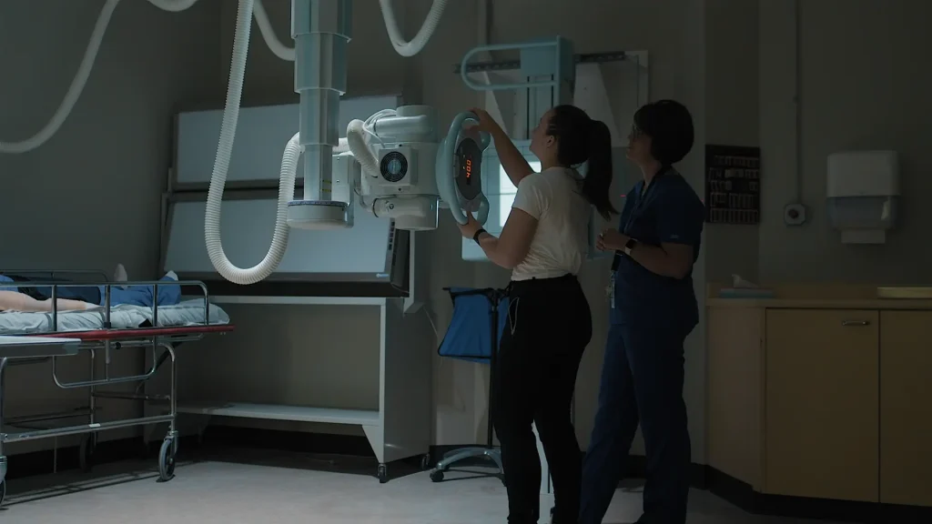 Two clinicians positioning a ceiling-mounted X-ray tube over a patient on a stretcher in a dim exam room