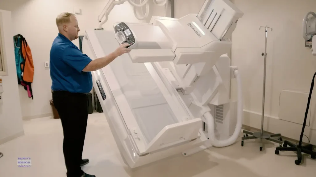 Technician adjusting the tilting table of a Shimadzu X1 R/F imaging system in an exam room