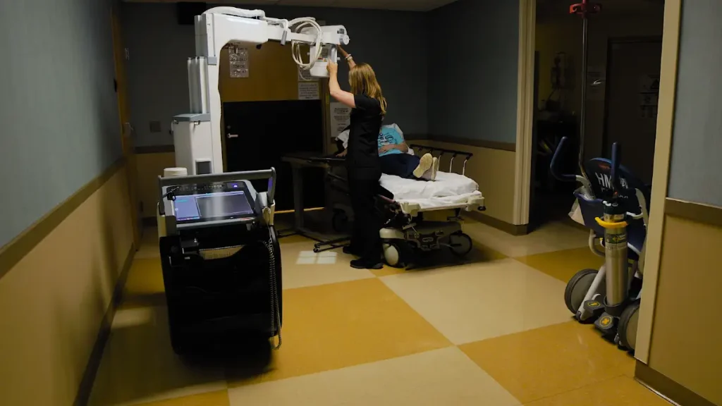 Radiologic technologist positioning a mobile X-ray unit over a patient on a hospital gurney in a hallway