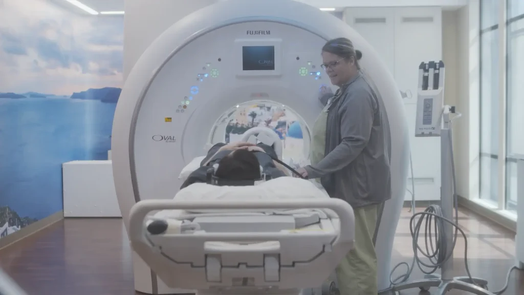 Technologist assisting a patient into a Fujifilm Oval MRI scanner in a room with a scenic wall mural