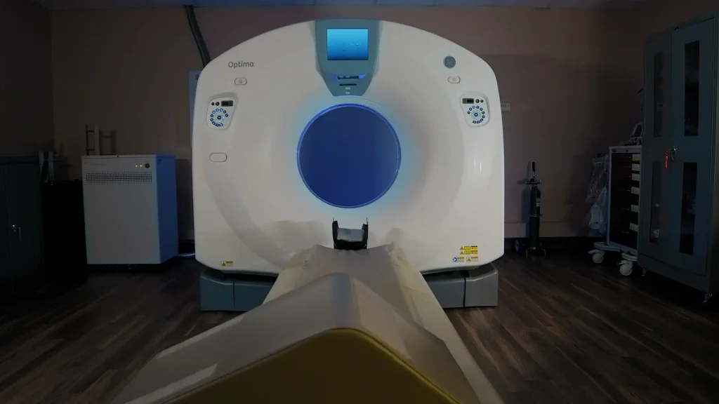 GE Optima CT scanner in an exam room, front view of the gantry with illuminated blue bore opening and patient table extended