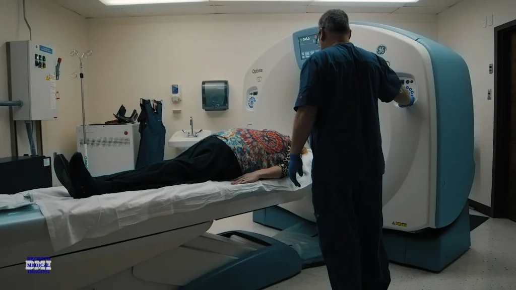 Technologist operating a GE Optima CT scanner console while a patient lies on the exam table
