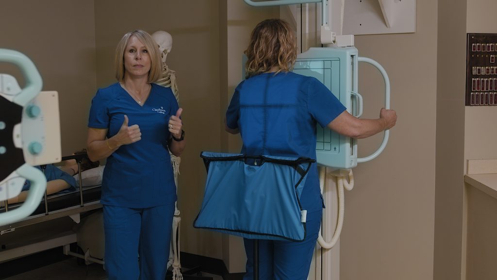 Two radiologic technology students practicing X-ray positioning, one giving a thumbs up while the other operates the detector panel