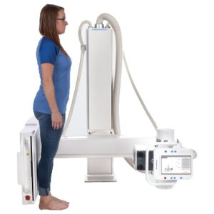Side view of a patient standing between the Essentia SA's wall-mounted detector stand and overhead tube column