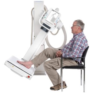 Patient seated with foot resting on the Essentia SA table detector for an extremity X-ray, with the tube head positioned overhead