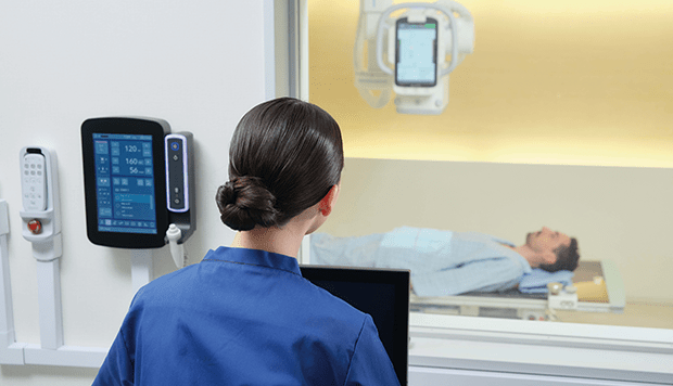Technologist operating the RADspeed Pro SR5 wall-mounted exposure console while a patient is positioned on the table in the exam room
