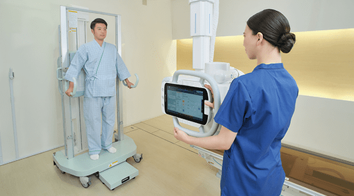 Technologist using a wireless tablet console to position a patient at the wall bucky stand on the RADspeed Pro SR5 system