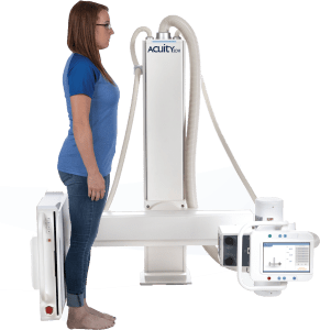 Acuity SDR+ radiographic system with a patient standing at the wall-mounted detector, showing the overhead tube stand and touchscreen control arm