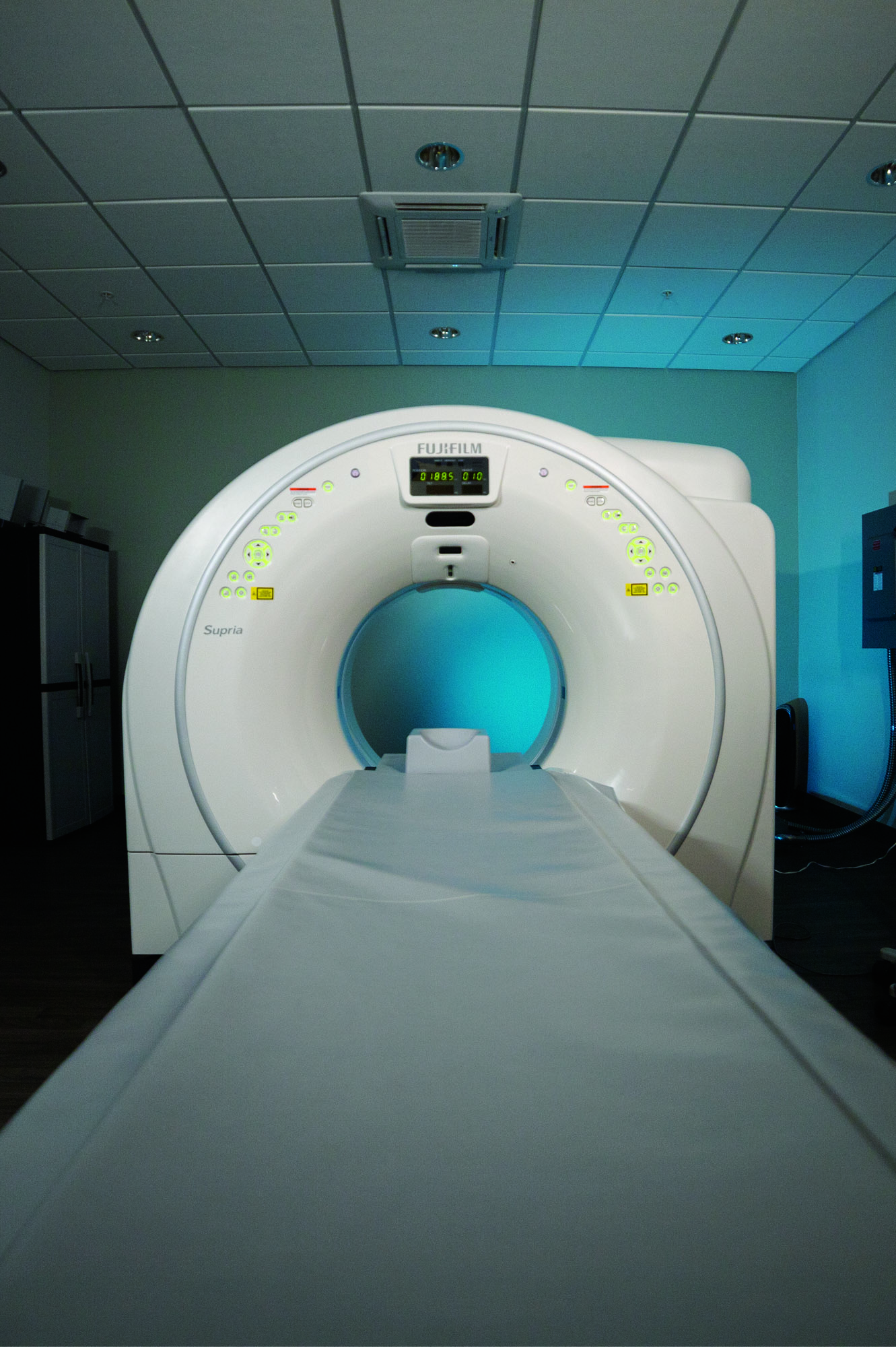Fujifilm Supria CT scanner gantry with patient table in exam room, teal ambient lighting