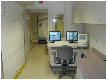 Mobile CT scanner control room with monitors and a chair inside a trailer, viewing into the scan room