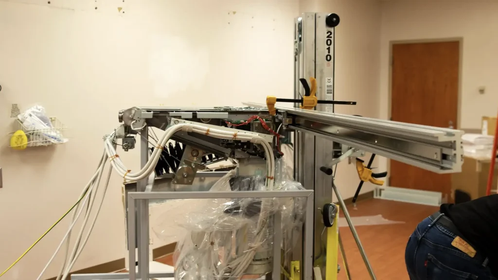 Technician servicing the open gantry of a CT or MRI scanner during installation, with an equipment lift in the room