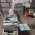 Agfa mobile X-ray unit with tube arm extended over touchscreen console displaying AGFA home screen in warehouse