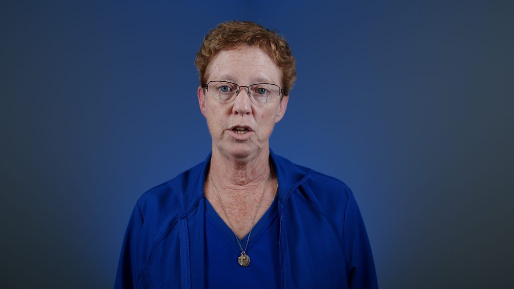Portrait of a woman with short red hair and glasses speaking against a blue background