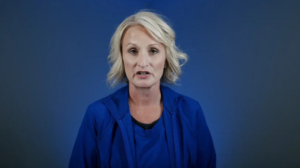 Portrait of a blonde woman in blue scrubs speaking against a blue background