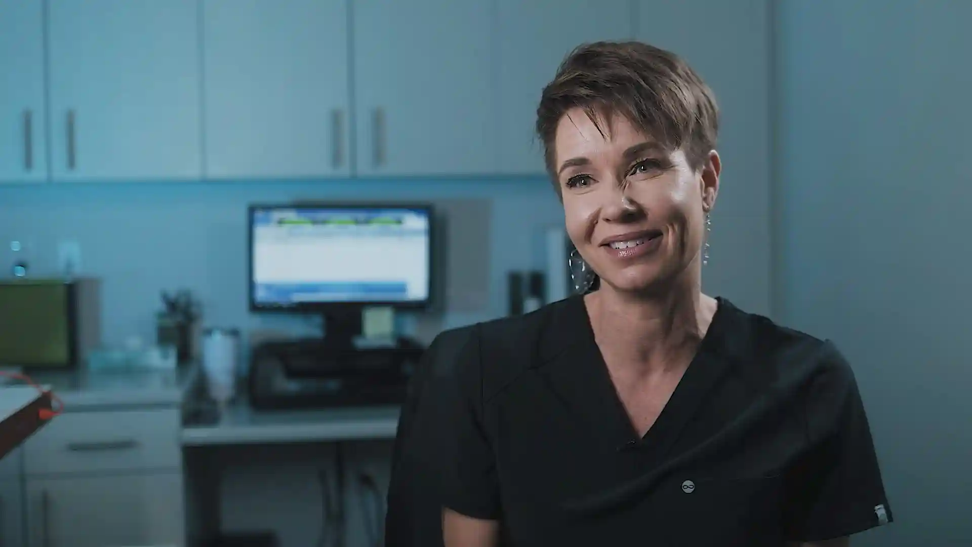 Smiling female technologist in dark scrubs in an exam room with a computer workstation behind her