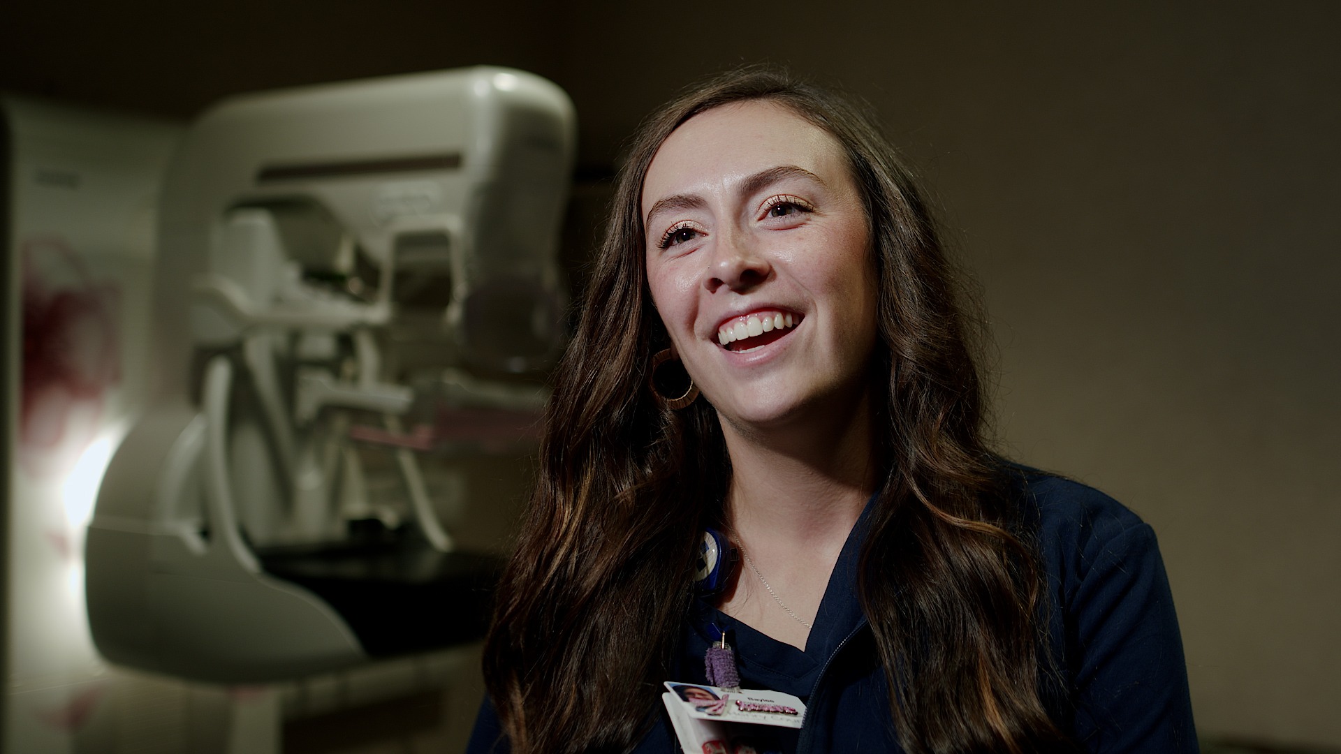 Smiling female technologist in navy scrubs with a mammography system blurred in the background