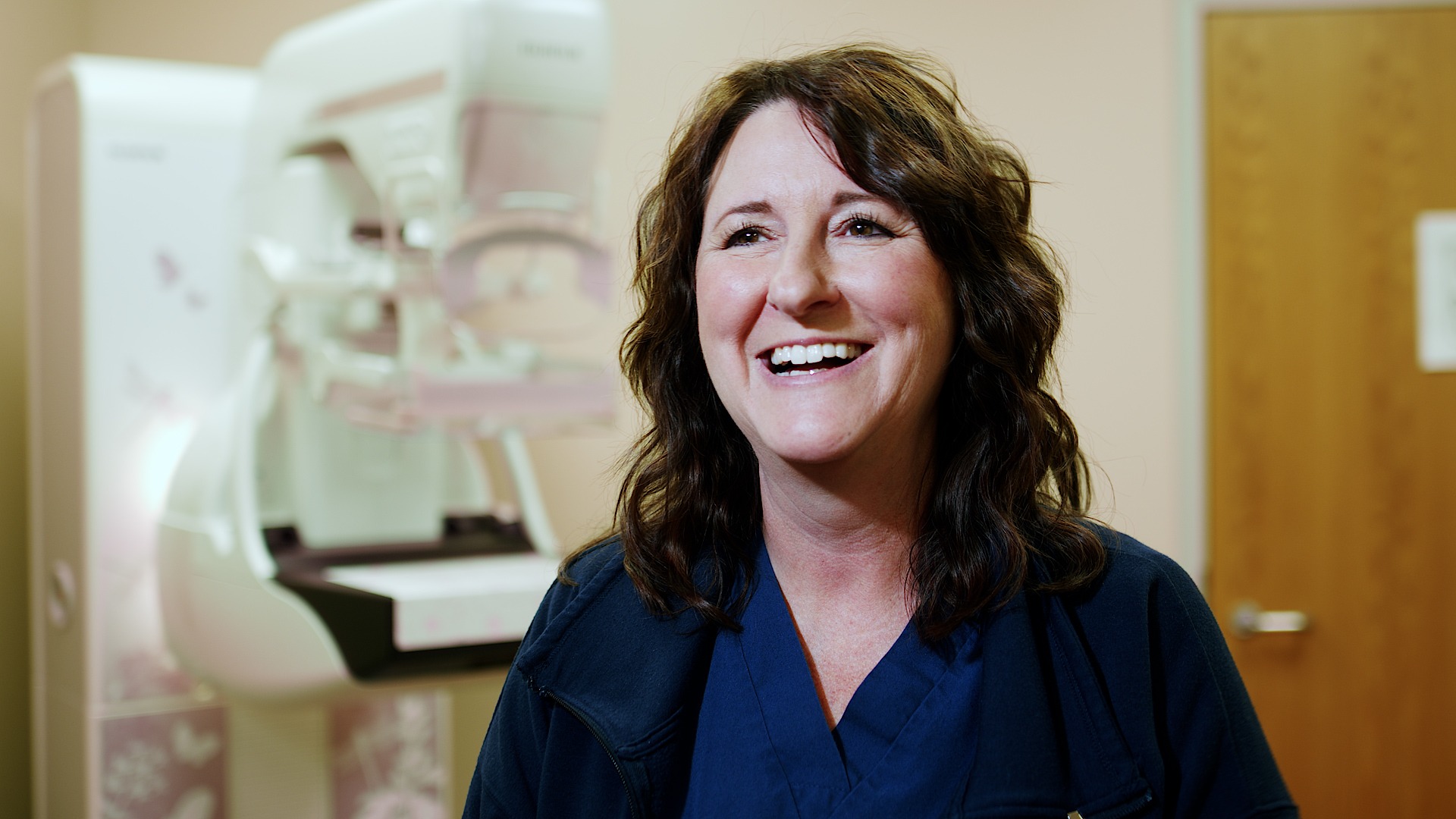 Smiling female technologist in navy scrubs standing beside a mammography system