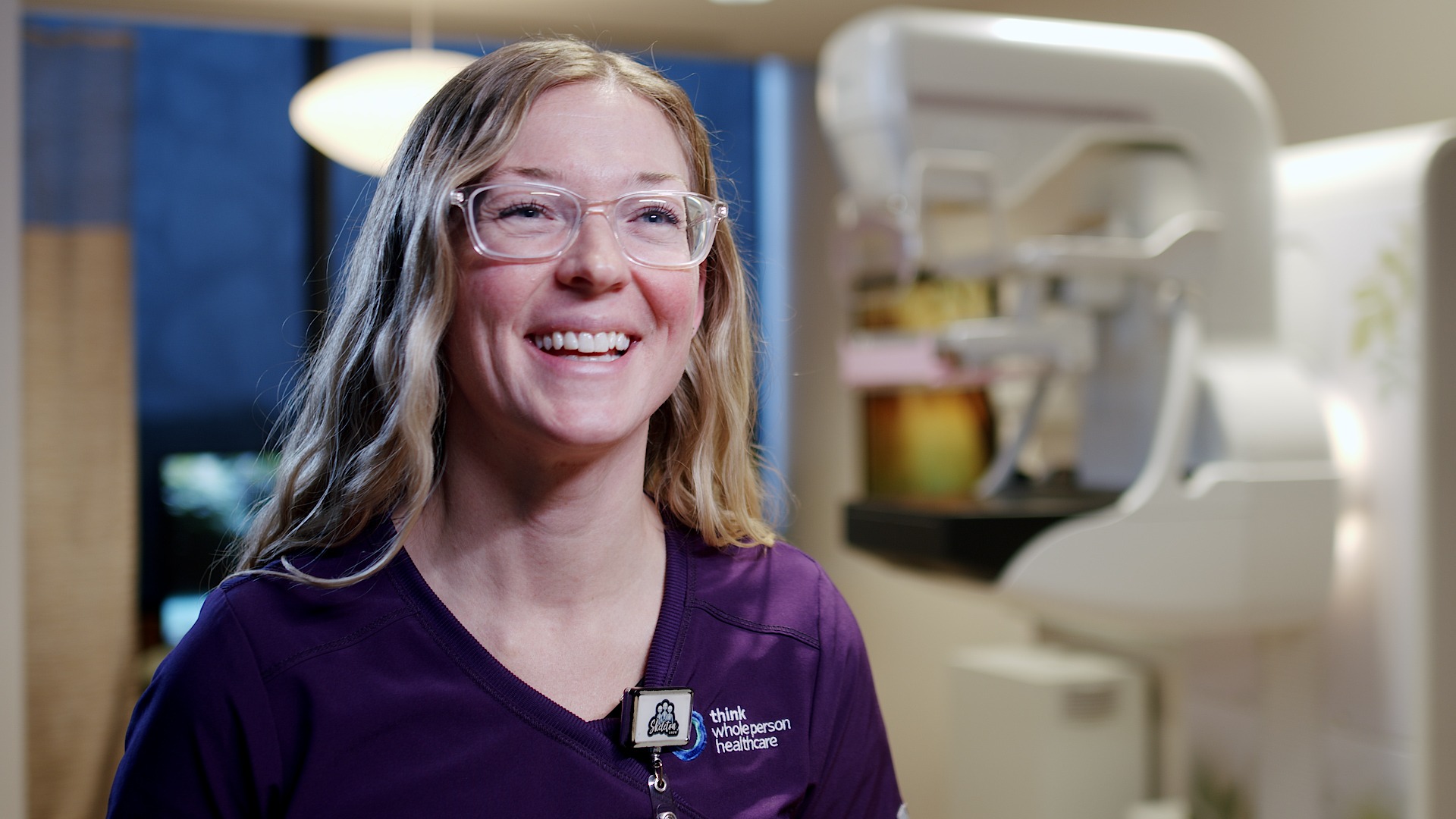 Smiling female technologist in purple scrubs with a mammography system blurred in the background