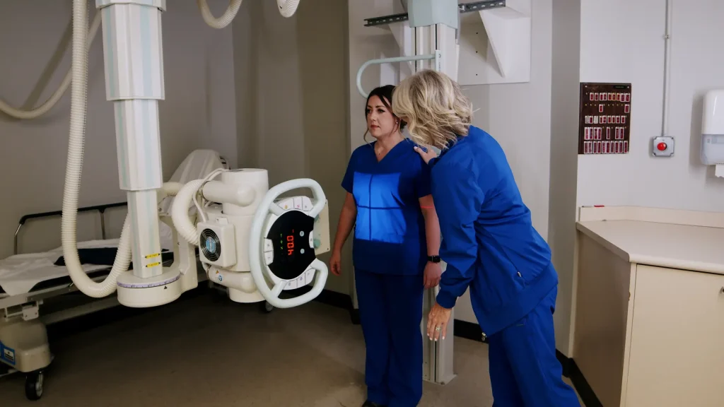 Two technologists positioning a patient against a wall-mounted digital X-ray detector for an abdomen exam