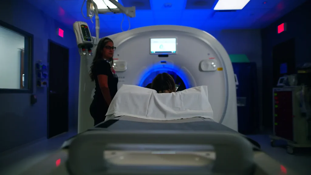 Video still of a technologist and patient at a CT scanner control area with blue ambient lighting