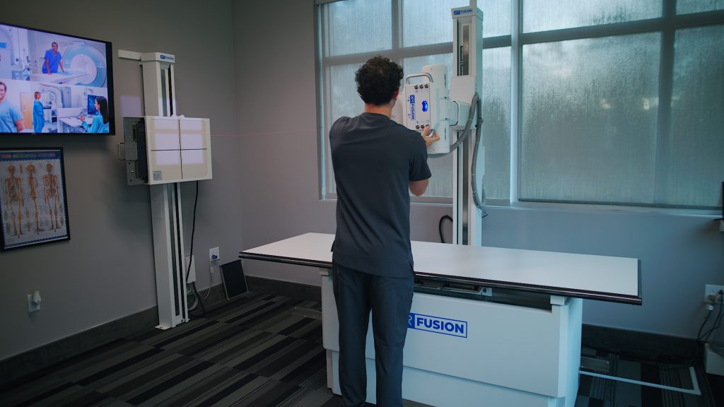 Technologist reviewing X-ray images on a wall-mounted monitor next to an XR Fusion imaging system