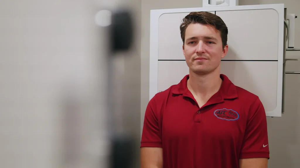 Man in a red polo shirt standing in front of a wall-mounted X-ray detector panel in an exam room