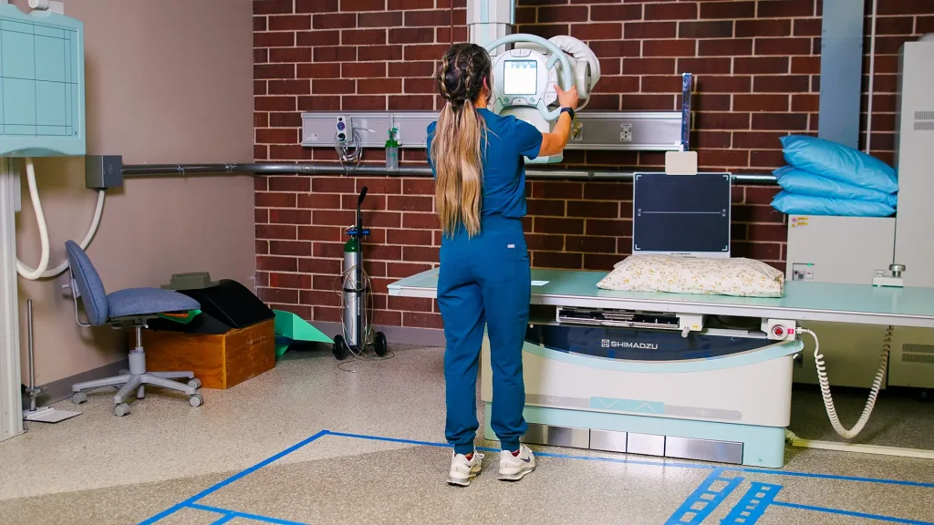 Technologist positioning a Shimadzu X-ray system's tube head over an exam table in a treatment room