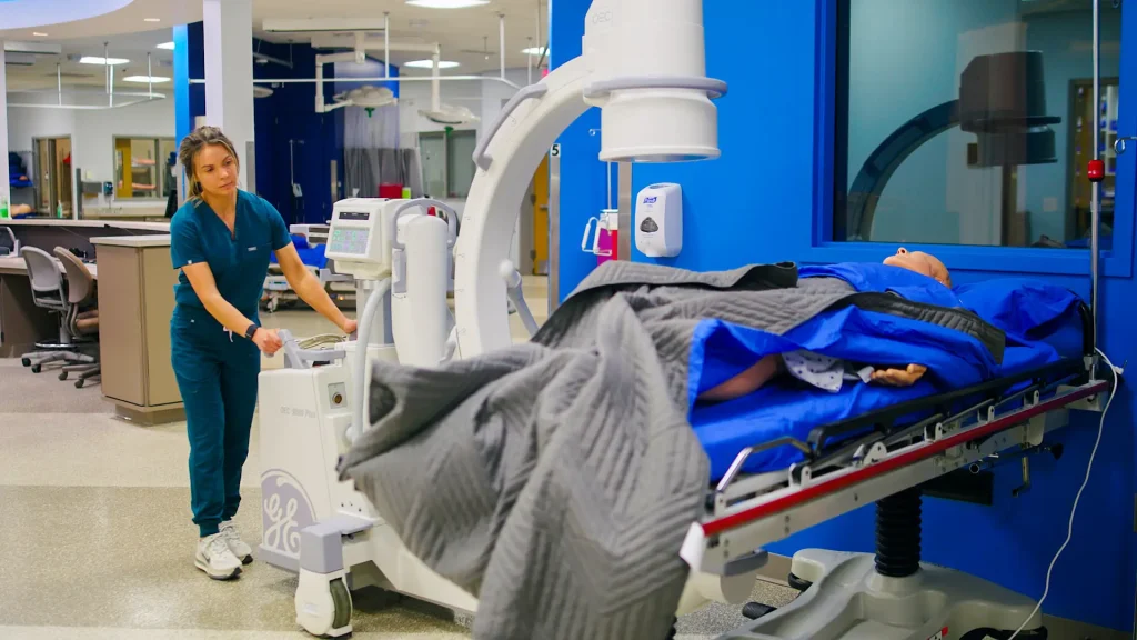 Technologist pushing a GE mobile C-arm X-ray unit toward a patient bed in a simulation lab